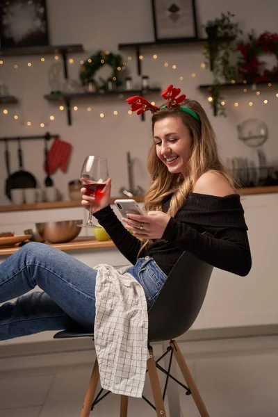 Ευτυχισμένη γυναίκα με ποτήρι κρασιού και τηλέφωνο κάθεται στην κουζίνα με φλεγόμενες γιρλάντες. — Φωτογραφία Αρχείου