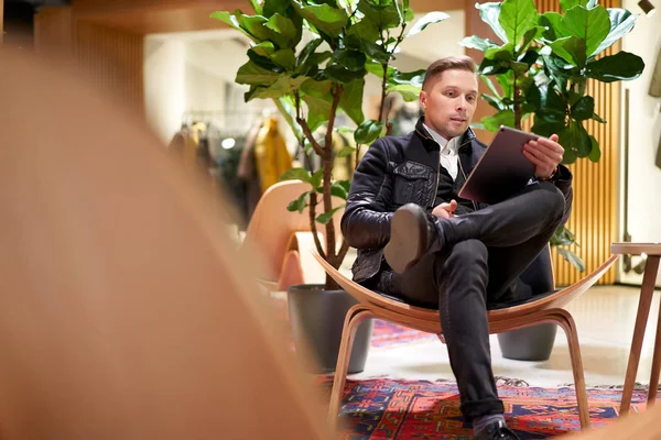 Giovane in giacca di pelle con tablet in mano seduto sulla sedia in negozio contro la pianta verde — Foto Stock