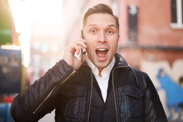 Překvapený muž mluví po telefonu stojí v blízkosti moderní budovy odpoledne. — Stock fotografie
