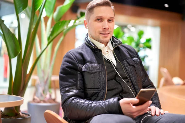 Brrunet macho com smartphone olhando para a câmera sentada na cadeira na loja contra planta verde — Fotografia de Stock