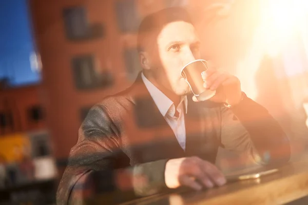 Молодий чоловік п'є зі скла, сидячи в кафе, відображаючи будівлю — стокове фото