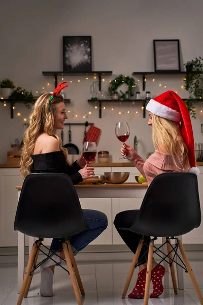 Noel Baba şapkalı ve geyik boynuzlu iki genç kadını Noel için mutfakta şarap kadehleriyle şenlik masasına oturtun. — Stok fotoğraf
