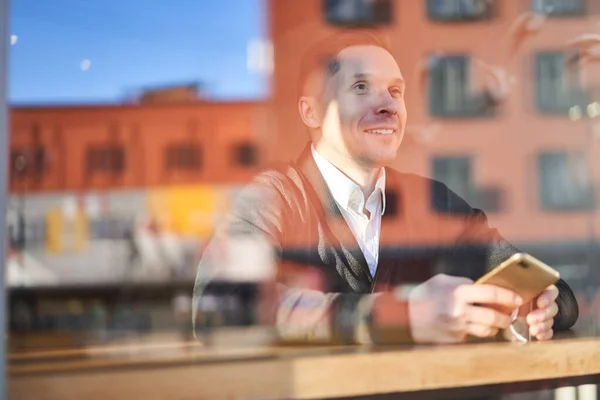 Smiling male with phone in his hand sitting in cafe, building reflection in glass