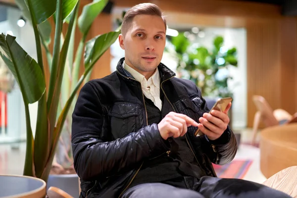 Muž v kožené bundě s chytrým telefonem dívá na kameru, zatímco sedí na židli v obchodě proti zelené rostliny — Stock fotografie