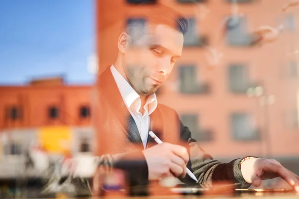 Hombre freelancer con teléfono y tableta sentado en la cafetería, reflexión edificio —  Fotos de Stock