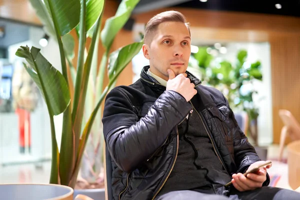 Hombre en chaqueta de cuero con teléfono inteligente mirando a un lado mientras está sentado en la silla en la tienda contra la planta verde —  Fotos de Stock