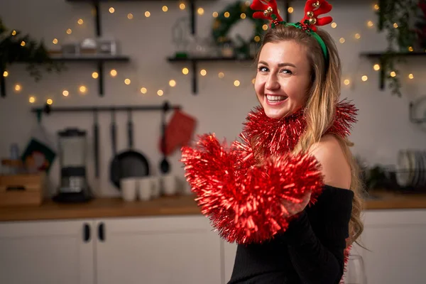 Glada kvinna med hjärta gjord av tinsel i händerna stående i köket under julen. — Stockfoto
