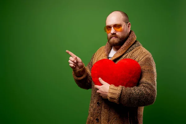 Komoly férfi rózsaszín szemüvegben tartja játék szív, miközben áll az üres zöld háttér stúdió — Stock Fotó