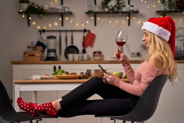 Молода жінка в капелюсі Санта з келихом вина і телефоном в руці сидить на кухні . — стокове фото