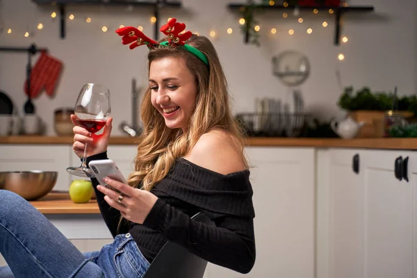 Szczęśliwa kobieta z kieliszkiem do wina i telefonem w ręku siedzi w kuchni na Nowy Rok — Zdjęcie stockowe