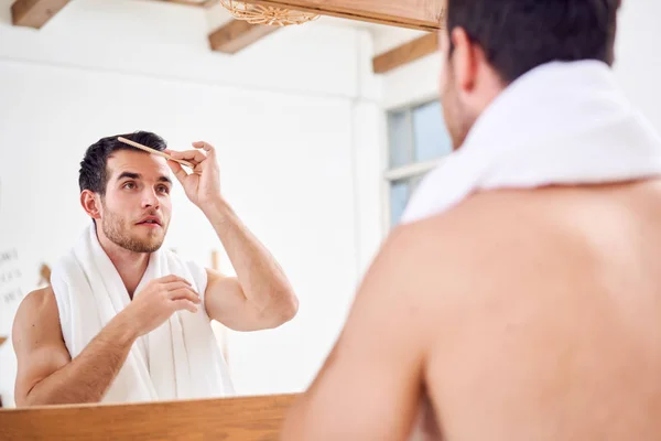 Unshaven man combing his hair while standing with white towel on neck near mirror in bathroom — Stock Photo, Image