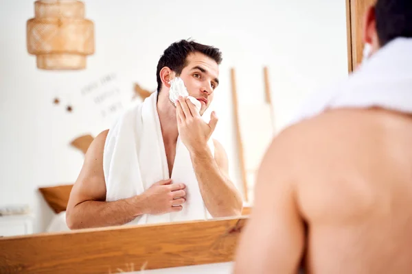 Młody brunet człowiek rozmazuje twarz z golenie pianka podczas stojąc w kąpieli w pobliżu lustro — Zdjęcie stockowe