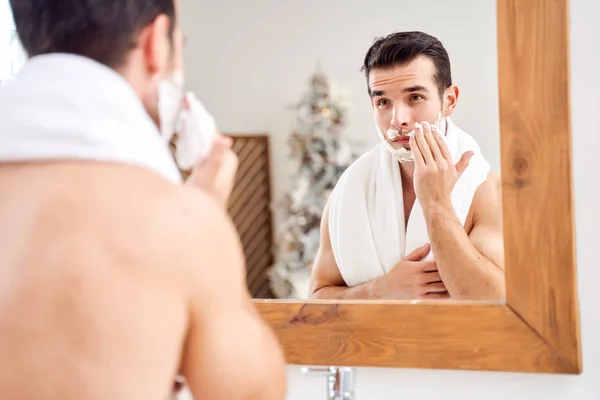 Brunet with towel on shoulders spreads face with shaving foam while standing in bath near mirror — Stock Photo, Image