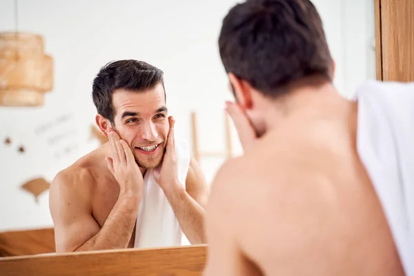 Young sports man lathers his face and stands with towel on his shoulders in front of bathroom mirror — 스톡 사진