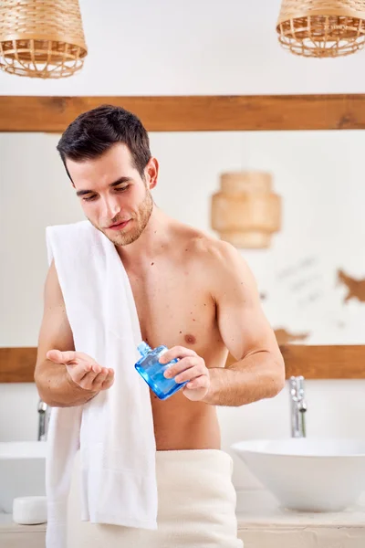 Brunet man with towel on his shoulders pours lotion into his hand stands in front of bathroom mirror — 스톡 사진