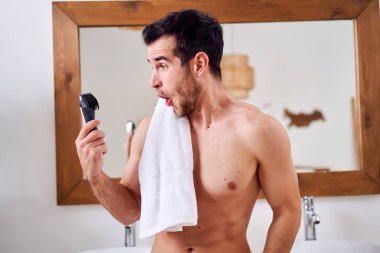 Elinde jiletle omzunda havluyla banyoda aynanın yanında duran genç bir adam.