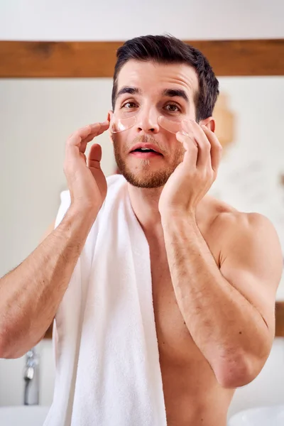 Brunet man applies transparent patches under his eyes while standing in bath opposite mirror — Stock Photo, Image