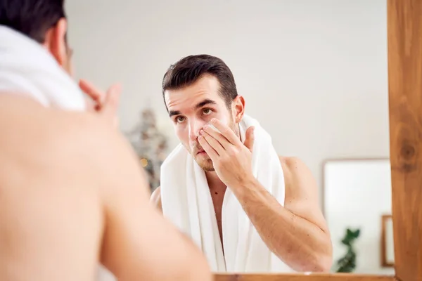 Young brunet man smears cream on face while standing near mirror in bath — 스톡 사진