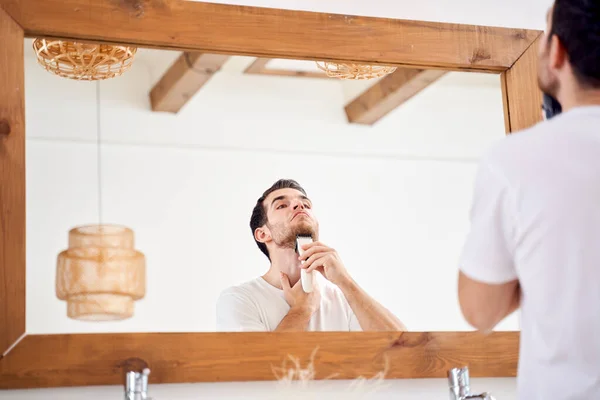 Брюне мужчина в белой футболке бреется, стоя возле зеркала в ванной — стоковое фото