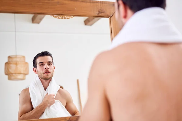 Young man with white towel on shoulder stands near mirror in bath at morning.