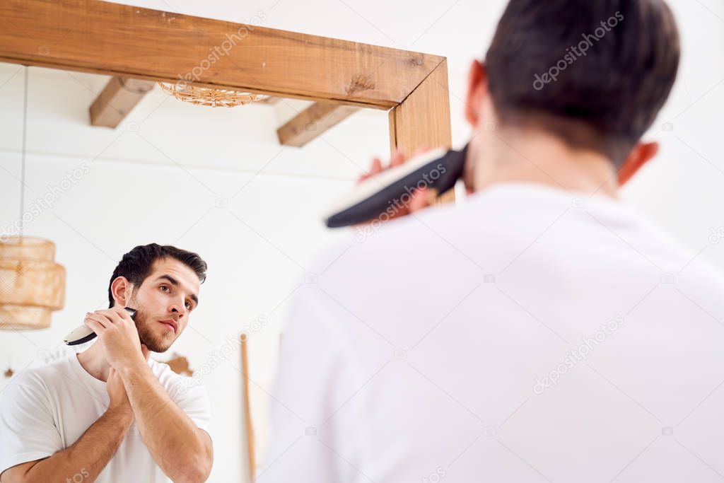 Close up brunet male in white T-shirt shaves while standing near mirror in bath