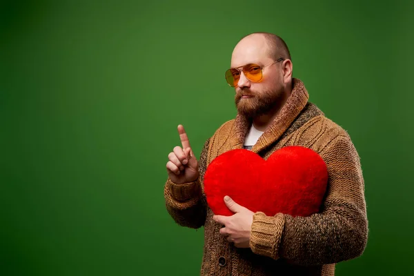 Férfi rózsaszín szemüvegben tartja játék szív, miközben áll az üres zöld háttér stúdió — Stock Fotó