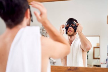 Yüzünde siyah maske olan genç bir adam banyoda aynanın karşısında duruyor.