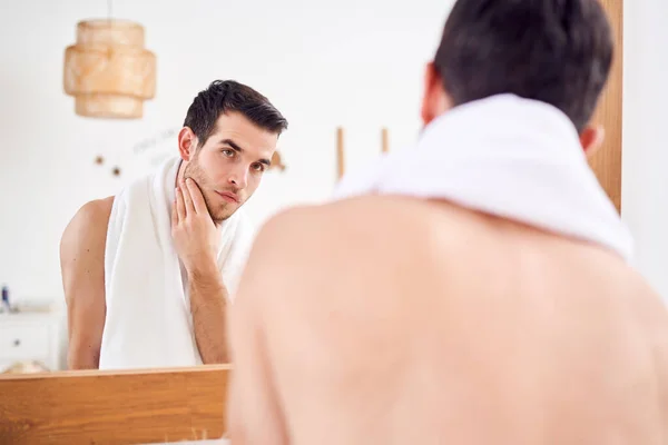 Unshaven naked young man with white towel on his neck standing near mirror in bathroom — Stock Photo, Image