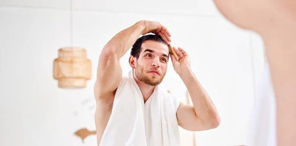 Young unshaven man combing hair standing near mirror in bathroom — Stock Photo, Image