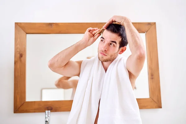 Brunet combing his hair while standing by mirror in bath — Stock Photo, Image