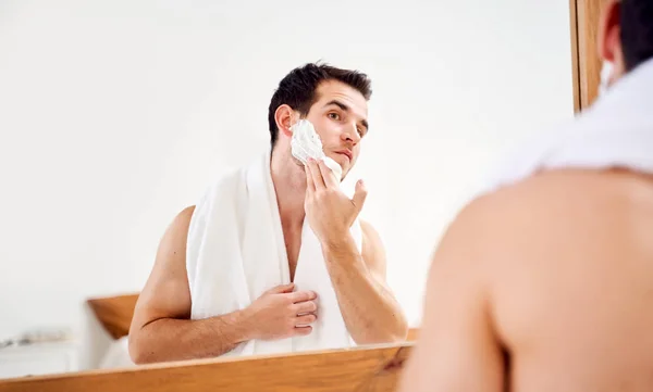 Hombre joven unta la cara con espuma de afeitar mientras está de pie en el baño cerca del espejo —  Fotos de Stock