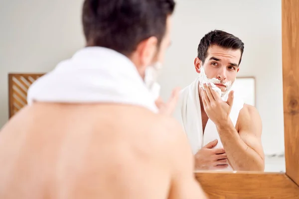 Young man with foam on beard near mirror in morning — Stock Photo, Image