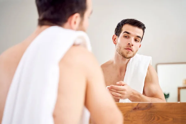 Brunet man spreads his face with white cream while standing in front of mirror in bath — Stock Photo, Image