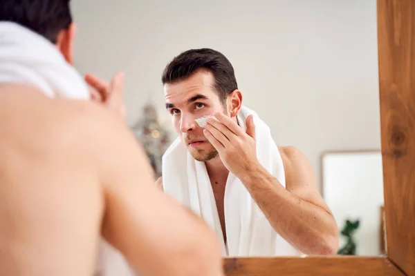 Young brunet smears cream on face while standing near mirror in bath — Stock Photo, Image