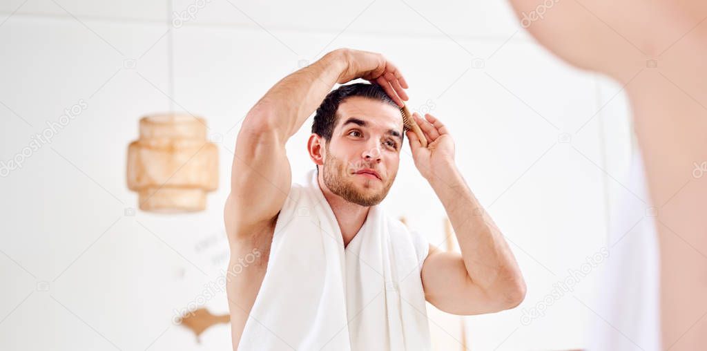 Young unshaven man combing hair standing near mirror in bathroom