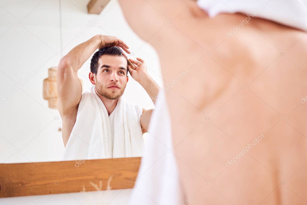 Man combing his hair while standing with white towel on neck near mirror in bathroom