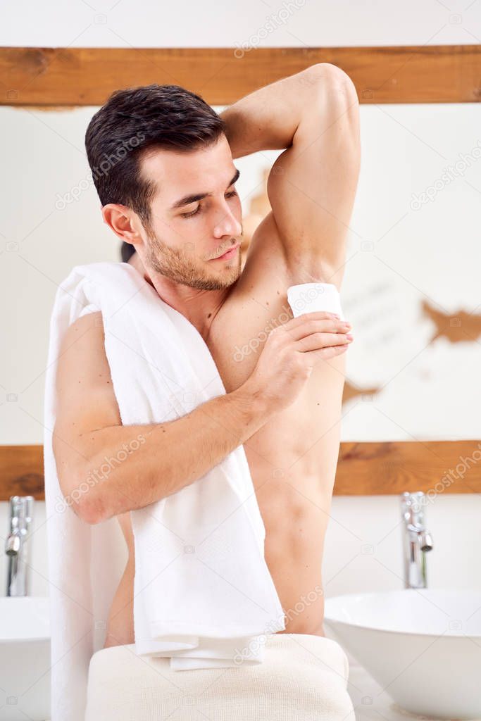Brunet male applies deodorant to armpits while standing in bath opposite mirror