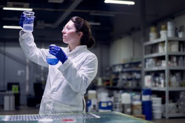 Young lab woman in glasses and white coat with flask with blue liquid in her hands checks quality