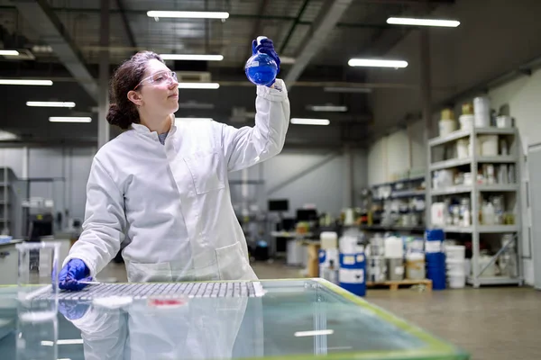 Mulher morena laboratório em jaleco branco com frasco com líquido azul em suas mãos verifica a qualidade — Fotografia de Stock