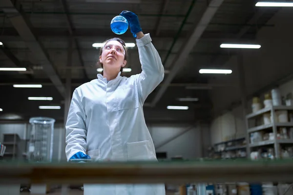 Young lab woman in white coat with flask with blue liquid in her hands checks quality on defocused background — Stock Photo, Image