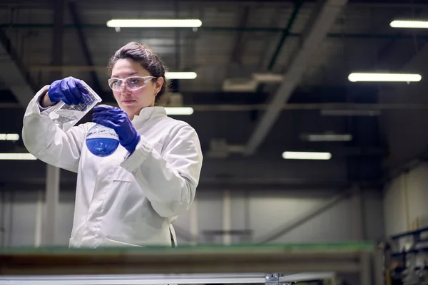 Молодая лаборантка в очках и белом халате с фляжкой с голубой жидкостью в руках проверяет качество — стоковое фото