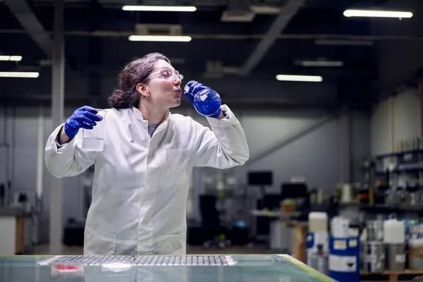 Молодая лаборантка в очках и белом халате с экспериментальной колбой с голубой жидкостью в руках проводит эксперименты — стоковое фото