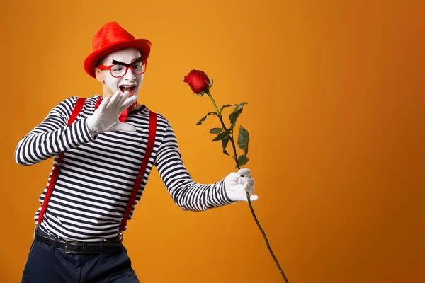 Vicces pantomim férfi fehér arc piros kalap és csíkos póló tartó rózsa elszigetelt üres narancs háttér — Stock Fotó