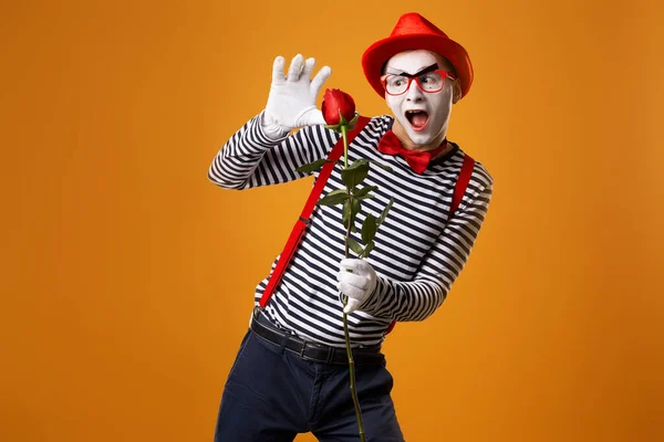 Rolig mimare med vitt ansikte i röd hatt och randig t-shirt innehav ros isolerad på tom orange bakgrund — Stockfoto