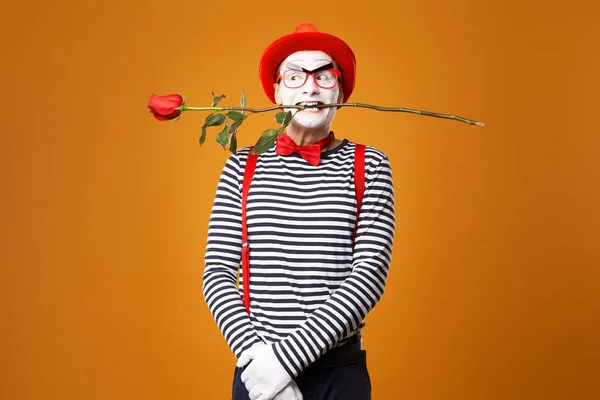Mime with white face in red hat and striped T-shirt holding rose in his mouth on blank orange background — 스톡 사진