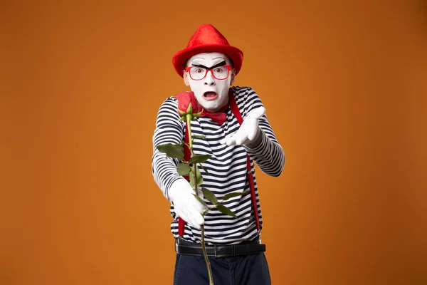 Triste mime homem em chapéu vermelho e colete com rosa nas mãos sobre fundo laranja — Fotografia de Stock