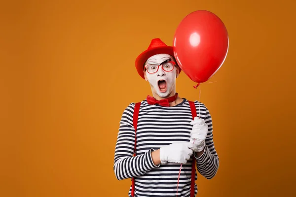 Překvapený mime muž v vestě a červený klobouk s balónem v ruce na oranžovém pozadí — Stock fotografie