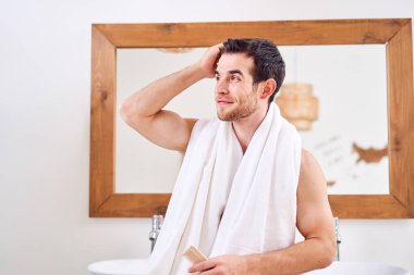 Boynunda beyaz havlu olan genç adam aynanın karşısında duruyor.