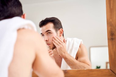 Genç esmer yüzünü köpürüyor ve omuzlarında havluyla banyo aynasının önünde duruyor.