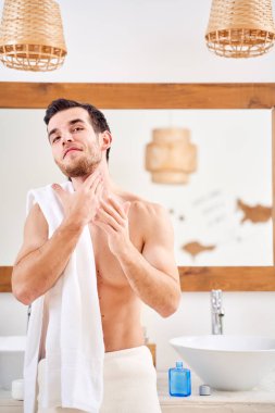 Genç adam banyo aynasının önünde omuzlarında havluyla duruyor.